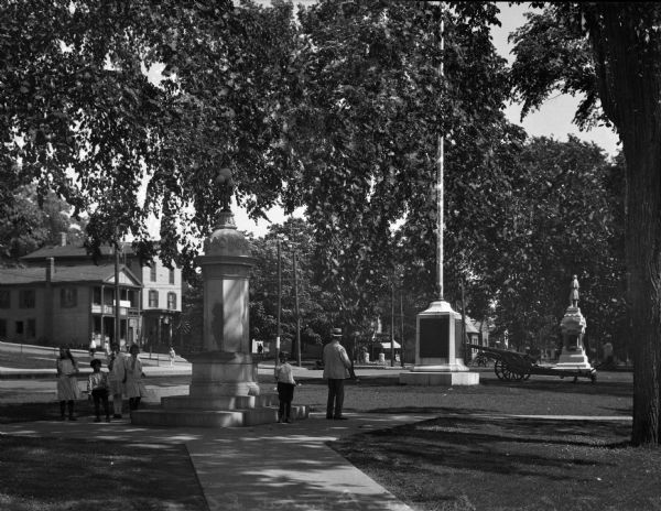 A view of five children eating ice cream and a man standing in a park with a number of monuments including a cannon, a flagpole with a stone base and a statue.