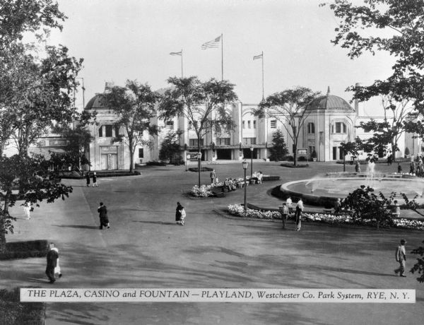 A view of Playland, an amusement center with a fountain, a plaza and a casino.  Pedestrians stroll across the plaza and three flags fly above the sprawling building complex's main entrance.  Published by Westchester Company.