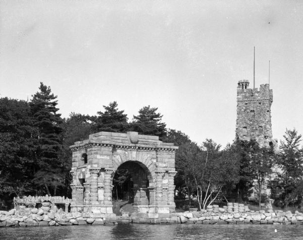 A stone arch and a stone tower standing among young trees overlooking the water. Loose stone blocks form the shoreline.