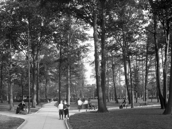 A view of Black Oak Park featuring two boys posing for the picture on a paved path through the tree dotted park. Other park-goers relax in the background on benches.