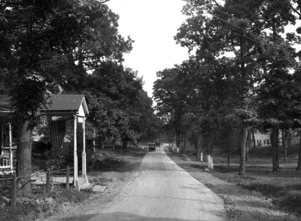 The entrance to Mountain Lake Park as seen from the tree-lined dirt roadway.  Two cars are visible in the distance further down the road and a covered porch entrance with a flag abuts the roadway.