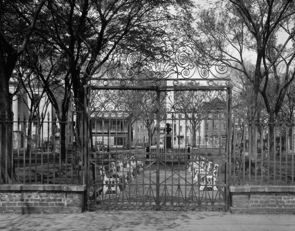 A view of an ornate metal gateway to a city park. Trees, facing rows of park benches and a bust on a pedestal can be seen through the gate.