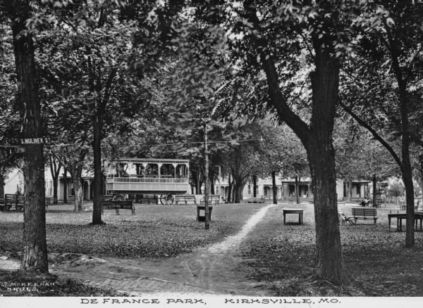 A view of DeFrance Park from S. Mulinex street featuring a dirt path, large trees, park benches and tables a large decorated cart and gazebos in the background.  Published by S. McKeehan Drugs.