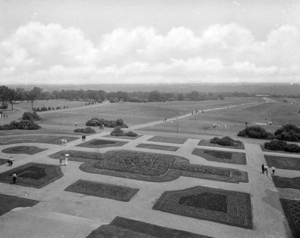 An elevated view of Siloam Gardens, a formal french garden, and the open countryside beyond. Pedestrians are strolling on the manicured and symmetrical paths of the garden and also about the grounds in the distance.