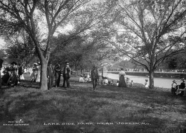 A view of Lakeside park showing parkgoers relaxing in the shade of the trees and the lake spanned by a footbridge in the background.