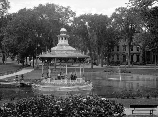 A view of an ornate gazebo set in a pond.  The pond has a fountain spray and tree-lined paved paths of the park are visible in the background while a flower bush and a park bench sit in the foreground.