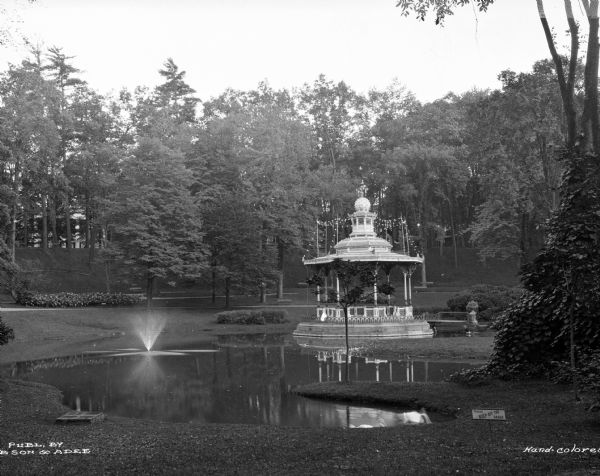 A view of an ornate gazebo strung with lights and set in a pond.  The pond has a fountain spray and is surrounded by grass and trees.  Published by bson [?] and Adee.