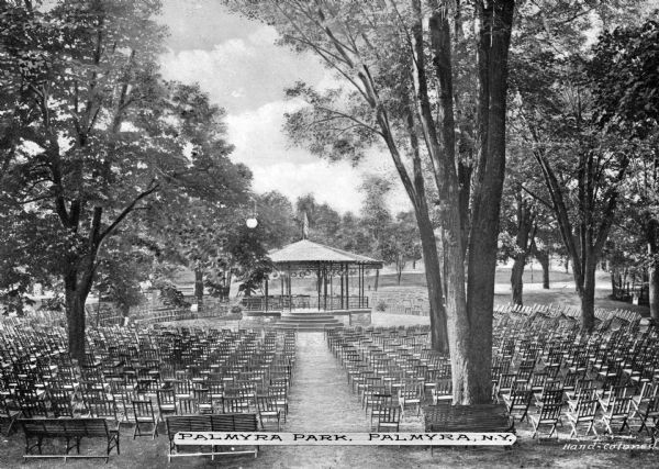 Rows of folding chairs set up in a park oriented to view a gazebo.  Tall trees partially shade the seating arrangement.