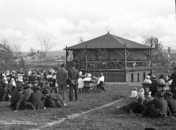 A view of a band stand in a park from behind a small crowd that has gathered to listen. The town is visible on the hisll in the distance.  Published by Kopp & Morris.