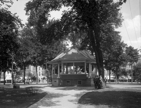View of a gazebo in Alcott Park.  A few men stand by a large tree that shades the gazebo and a street lined with shops is visible in the background through the other park trees.