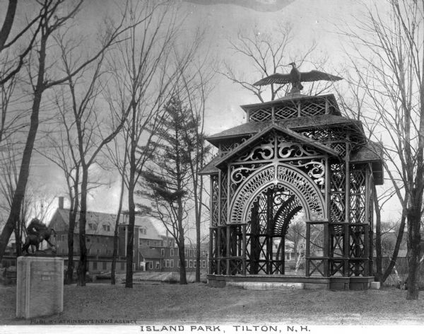 A view of Island Park and an elaborately carved open air pavilion surmounted by a large carved bird with outstretched wings.  A small sculpture of a figure on a horse sits on a pedestal nearby and a large building is visible in the background.  Published by Atkinson's News Agency.