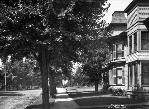 A view of the West Atwood Street residential district featuring a Victorian house with scalloped shingles and a turret. Many trees shade the street that stretches into the distance. Published by J.M. Miller.