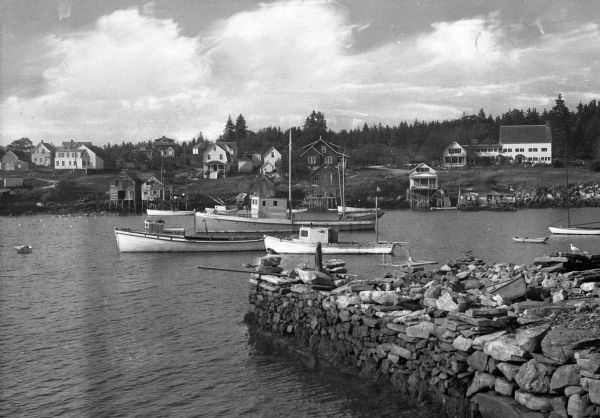 View across harbor filled with boats toward small wooden cottages along the far shoreline. A rock wall along the water's edge is in the foreground on the right.