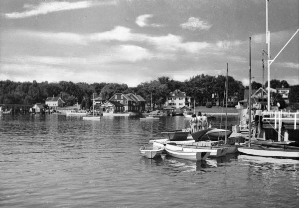 View across water towards piers, boats and people in a harbor. Dwellings and other buildings are on the far shoreline.
