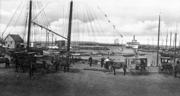 A view toward a wharf with horse-drawn vehicles in the foreground, and a steamship in the distance.