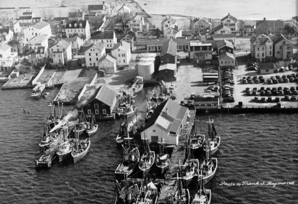 Aerial view of a harbor with a wharf in the foreground, and buildings and boathouses along the shoreline.