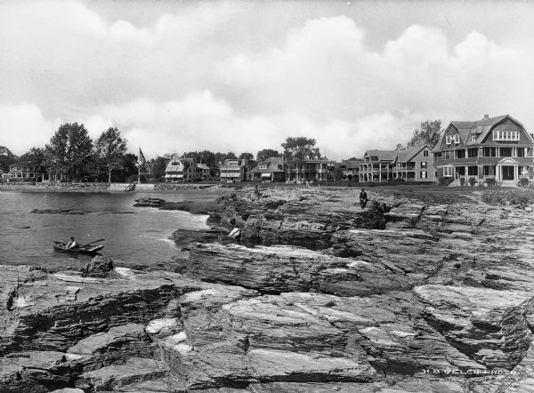 View along a rocky coastline toward resort hotels. People are sitting on the rocks, and a man is in a boat near the shoreline.