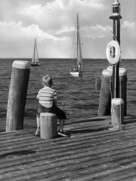 A view of a boy watching the sea from a pier. Two sailboats are in the water,