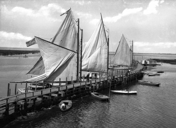 Elevated view of a long, curving pier on the bay with large sailboats with sails out on the left side, and smaller boats on the right.