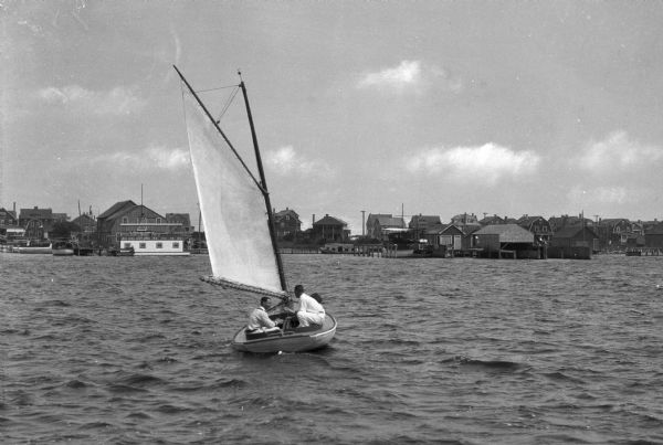 View across water toward people on a small sailboat on Barnegat Bay, and boathouses and buildings on the far shoreline.