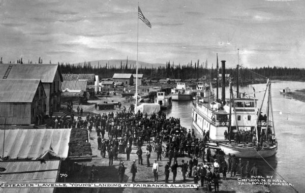 Elevated view of the steamboat "Lavelle Young" landing in Fairbanks as a crowd of people are looking on. Text on photograph reads: "Steamer 'Lavelle Young' landing at Fairbanks, Alaska." and "Publ. By Chisholm & Hall Fairbanks, Alaska."