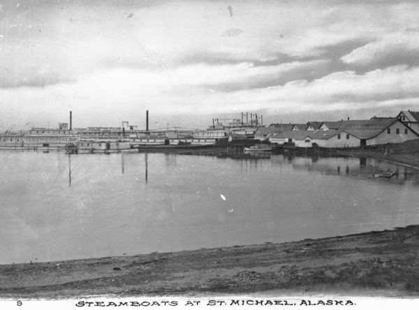 View over shoreline toward steamboats moored out in a harbor with buildings lining the far shoreline. Caption reads: "Steamboats at St. Michael, Alaska."