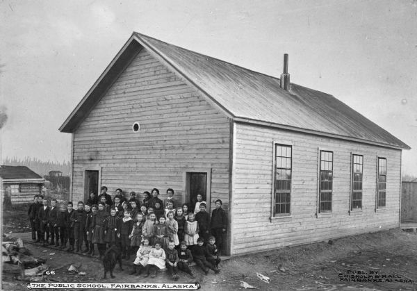 A public schoolhouse with a group of children and a dog posing in front. Text in lower left corner reads: "The Public School, Fairbanks Alaska."