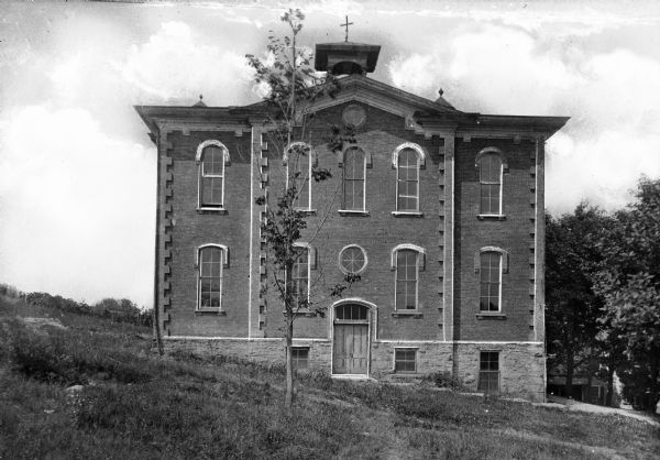 View of the exterior of an unidentified brick high school with a crucifix on top of the belltower on the roof.