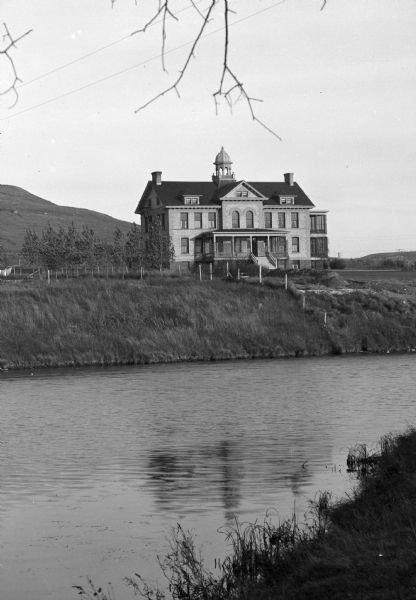 View from shoreline across water toward an unidentified school building with an ornate bell tower on the roof on the bank of a river or large pond. A hill is in the background on the left.