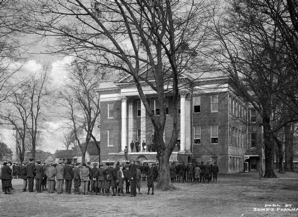 Exterior view of the boys high school building, with boys standing in lines in front of the building.