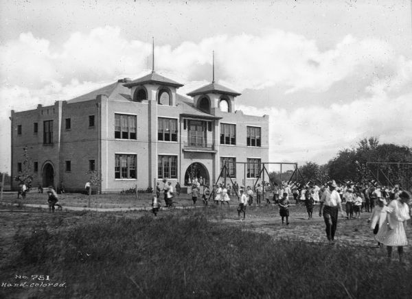 View of First Ward school with children playing on swings in a playground area in the foreground.