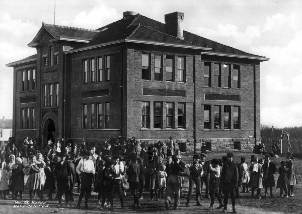 Exterior view of Jefferson School with a number of students congregating in a courtyard area in the foreground. Women are standing near the building with other children.