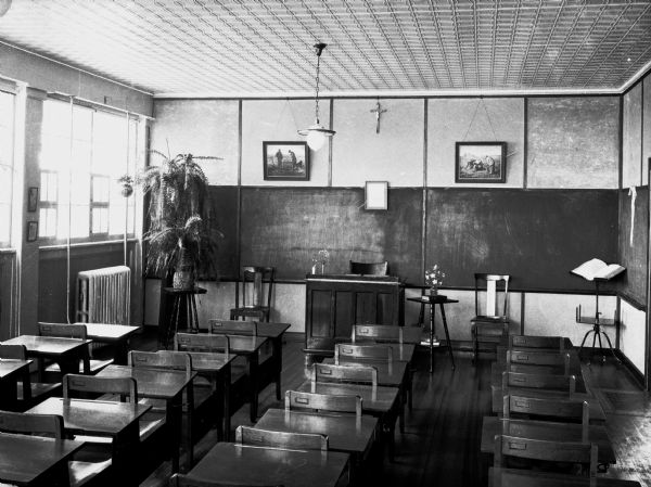 Slightly elevated interior view from the back of a classroom in Notre Dame Convent. There are rows of desks, windows along the left wall, a lamp hanging from the ceiling, and a crucifix on the wall at the front of the room.