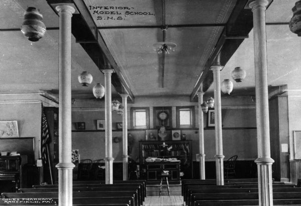Interior view from the back of the room of a model school, with rows of benches facing the elevated desk and bookshelf. There is a piano on the far left. Text reads: Interior - Model School, S.N.S."