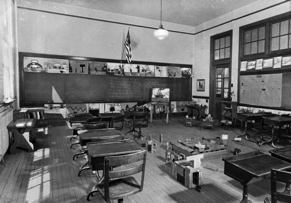 An interior view of a classroom in Bywood School. Desks ring the room, leaving a play area littered with blocks, and chalkboards and an American flag are on the wall.