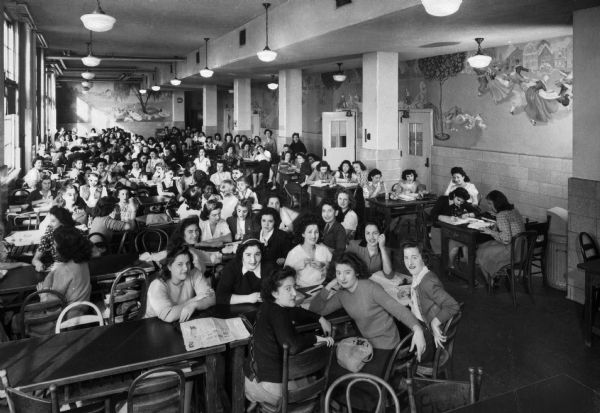 Elevated view of the lunch room of Washington Irving High School, designed by the architect C.B.J. Snyder (1860-1945) and built in 1913. Lights hang from the high ceiling, murals adorn the walls, and a number of girls are seated at the long tables.