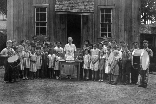 A large group of children posed outdoors at a birthday party for a teacher at Berry School. The teacher is at the center and is surrounded on both sides by children, the ones on both the far left and right hold large drums.