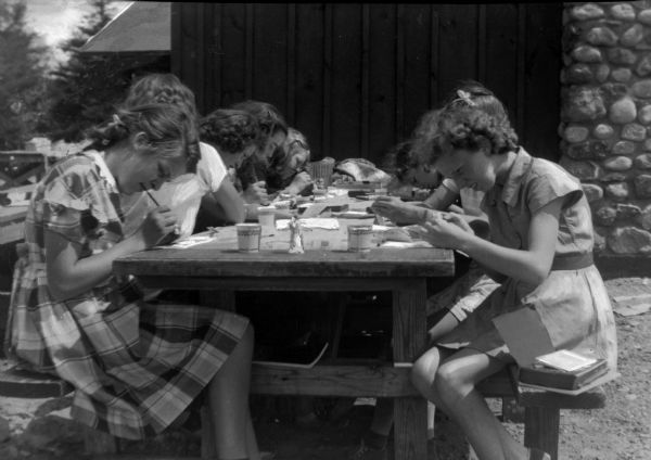 Girls sitting at a picnic table enjoy an outdoor craft class at Camp Alobregate. A building with a stone chimney is in the background.