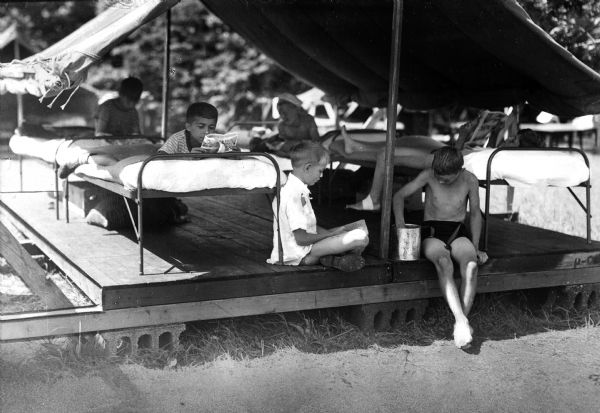 A view of boys reading and relaxing underneath a semi-permanent canvas-roofed tent on a platform at Camp Wanasquetta.