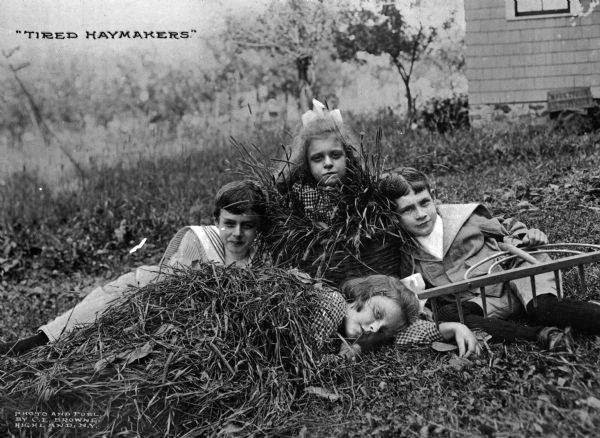 Four children pose for a photograph, one holding a large wooden rake, while playing in the hay. Photograph published by C. E. Browne. Text in upper left corner reads: "Tired Haymakers."