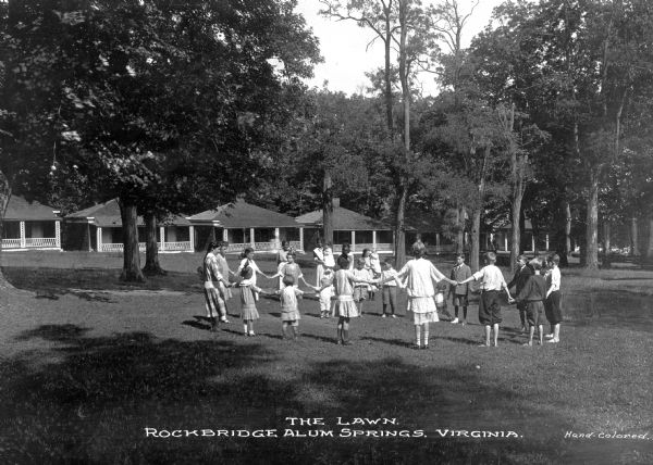 Women and children play on the lawn in front of the cabins of Rockbridge Alum Springs, a camp for children. The children and adults are holding hands in a circle and there are camp buildings visible in the background. Caption reads: "The Lawn. Rockbridge Alum Springs, Virginia."