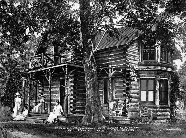 Edmund Gardner's log cabin, purchased by Gardner in 1874. A group of women are on the porch. Caption reads: "Comm. Gardner's Log Cabin at Ballas Island."
