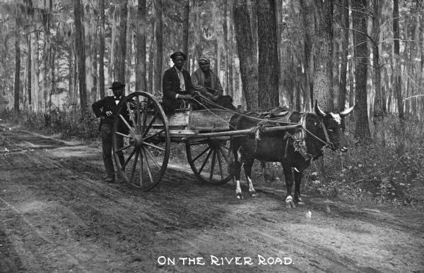 Three people pose in and around an ox cart on a country road. Caption reads: "On the River Road."