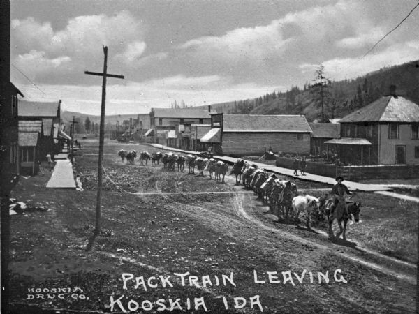 Slightly elevated view of a pack train leaving town. Caption reads: "Pack Train Leaving Kooskia Ida."
