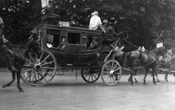 View toward the right side of an original 19th century stagecoach, called "Hiawatha," known for carrying passengers along the banks of the Delaware, the United States mail, and the business of the local Wells Fargo Express Agency. People are riding on top of the stagecoach, and other people are sitting inside. A man is riding a horse behind them on the left.