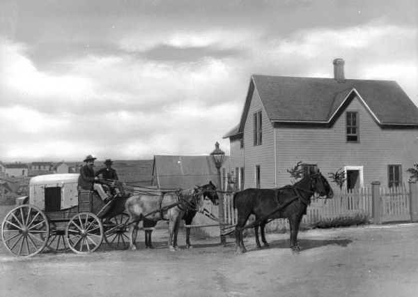 A team of four horses with two men on a stagecoach in front of a house.