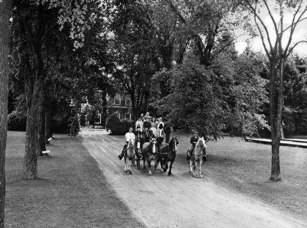 Slightly elevated view of a group of people on a horse-drawn coach, riding down a residential drive in Lenox. A large house is in the background behind trees. Two people riding on horseback flank the coach.