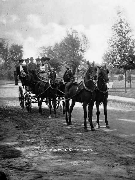 A horse-drawn carriage with a team of four horses with passengers passing through City Park. Caption reads: "View in City Park."