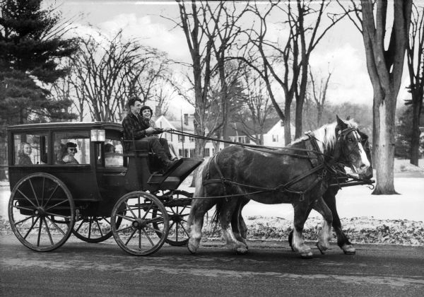 A group of men and women in a horse and coach on a country road with the Eastover resort estate in the background. Eastover resort estate was built in 1910 as a summer cottage for Harris Fahnestock and his family.