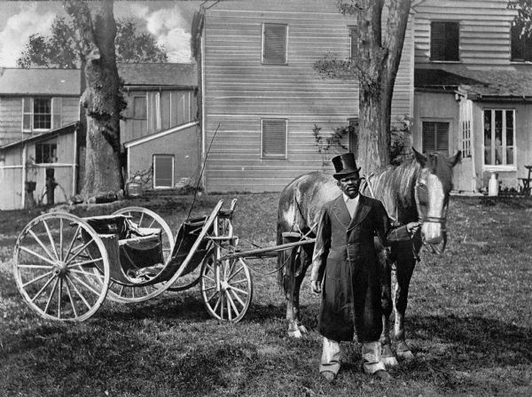 A man wearing a top-hat and a long coat is standing on the lawn of a house holding the reins of a horse pulling a three-wheeled chaise. In the background is a large dwelling.